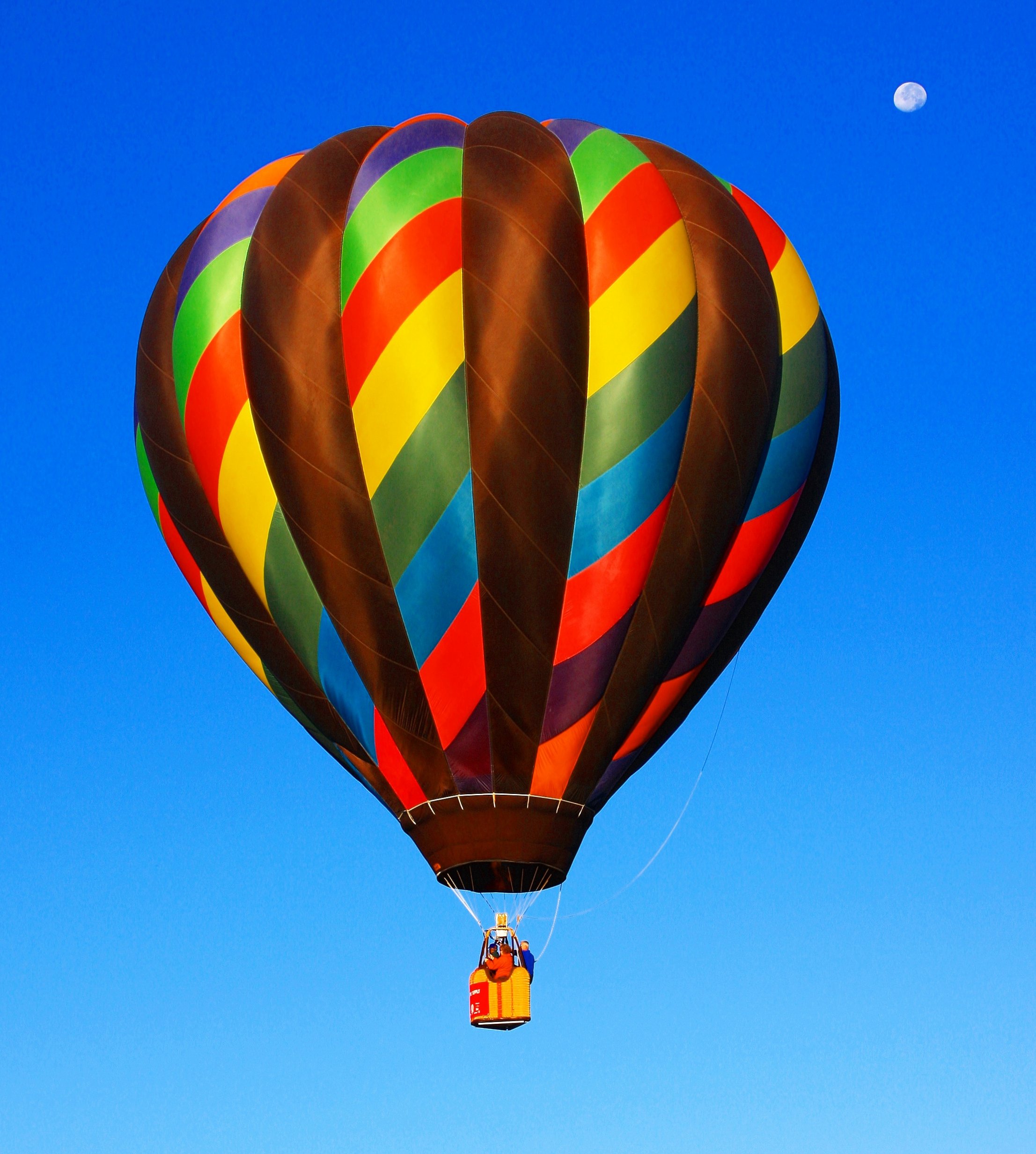 A striped hot air balloon floating in a blue sky, darker toward the top of the image. A small, nearly full moon is visible; the ground is not.